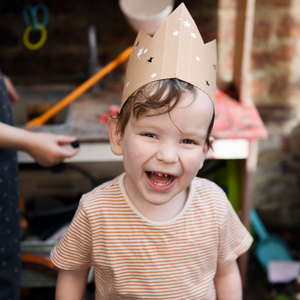 Young child with a big grinning smile, they are wearing a cardboard birthday crown that is covered in a white star pattern.