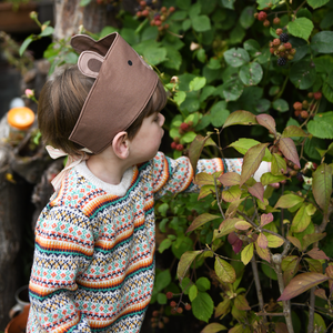 A young child wearing a knitted jumper and a bear dress up fabric crown picks blackberries from the bush.