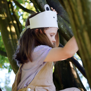 A young girl wearing a dress-up polar bear fabric crown and beige dungarees is climbing a tree.