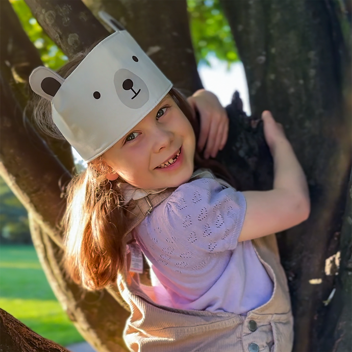 A young girl wearing a dress-up polar bear fabric crown and beige dungarees is climbing a tree.