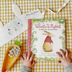 An illustrated copy of The Velveteen Rabbit book by Margery Williams and Sarah Massini lays on a yellow gingham table. A handmade bunny crown lays beside it, and a young child holds the book at the bottom.