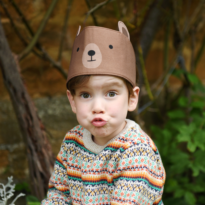 A young child wearing a knitted jumper and a bear dress up fabric crown sits outside pulling a funny face!