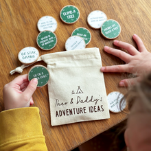 A child’s hands holding wooden adventure tokens over a wooden table. The tokens, featuring various outdoor activity suggestions, are scattered around a small canvas drawstring bag labelled "Theo & Daddy's Adventure Ideas."