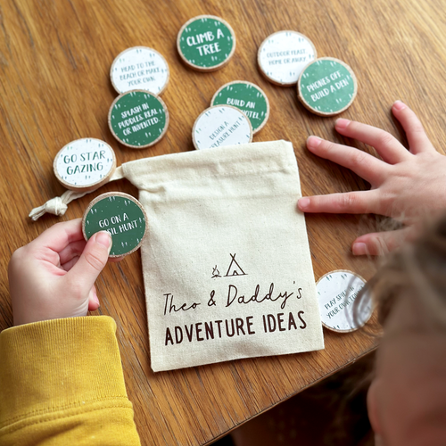 A child’s hands holding wooden adventure tokens over a wooden table. The tokens, featuring various outdoor activity suggestions, are scattered around a small canvas drawstring bag labelled 