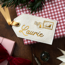 A personalised cream-colored felt luggage tag with gold foil embroidery reading 'Laurie' and a gold Christmas-themed stamp design. The tag is attached to a gold ribbon and is styled on top of a red and white gingham wrapped gift box, surrounded by festive props like pine branches, a pink box with a red ribbon, and a dark glass bottle on a wooden surface.