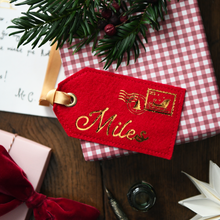 A red felt Christmas gift tag with gold embroidered script reading 'Miles' and a postage-style stamp motif, attached to a red-and-white gingham gift box on a wooden tabletop with holiday greenery and gift props around it.