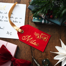 A red felt luggage tag with gold script reading 'Miles' and a gold postmark, laid on a wooden tabletop with gift boxes, greenery, and a letter nearby.
