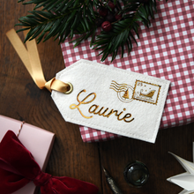 A personalised white felt luggage tag with gold embroidered script reading 'Laurie' and a gold postage stamp motif, attached to a gold ribbon and styled on top of a red-and-white gingham gift box with pine branches, a pink box with a red bow, and a dark wooden surface.