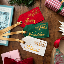 Three personalised felt Christmas postmark tags in red, green, and white with gold embroidered names and stamps, attached to gold ribbons, arranged on a wooden tabletop with wrapped gifts and holiday decor.
