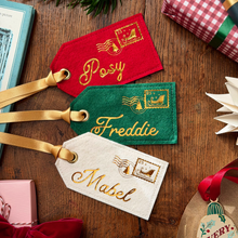 Three personalised felt luggage tags in red, green, and cream, each with a gold ribbon loop and gold embroidered postage stamp and name. The tags are arranged on a wooden tabletop with holiday gift props around them.