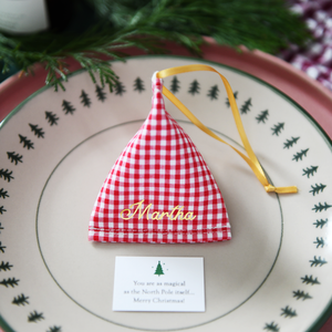 A red gingham fabric fortune cookie with gold personalised name “Martha” sits on a Christmas dinner plate, ready as a handmade festive place setting with a hidden Christmas wish card beneath.