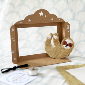 A handmade sloth finger puppet sits inside a tiny cardboard puppet theatre.