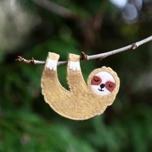 A handmade sloth finger puppet made from beige and white felt is hanging from a tree branch.