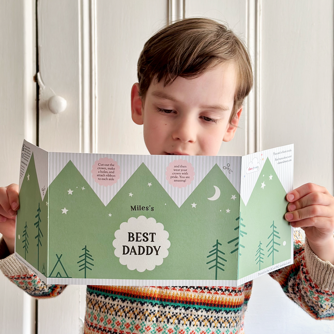 A young child wearing a knitted jumper holds a Father's Day card with an adventure crown design. The green crown has the words 'Miles Best Daddy' in the centre and a pattern of camping and tree illustrations. The card can be turned into a wearable crown for Father's Day.
