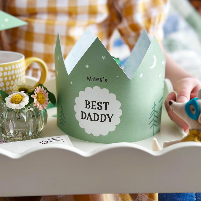 A personalised Best Dad crown with a green adventure design stands on a Father's Day breakfast tray that is being held by a young child. The wooden tray has a cup of tea, gifts, and keepsakes for Daddy. It's a lovely sentimental scene for Father's Day gifting.