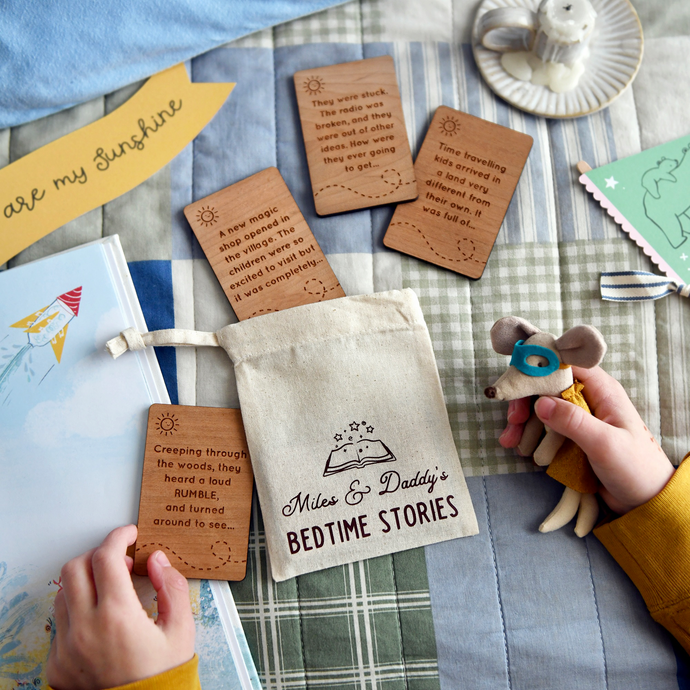 Personalised bedtime story set for dads and children featuring engraved wooden story prompt cards and a drawstring cotton bag reading 'Miles & Daddy’s Bedtime Stories', displayed on a patchwork quilt with a soft toy, picture book, and a young child holding one of the cards.