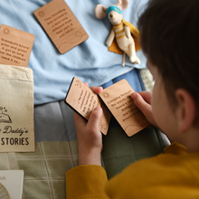 A young child is lying on a bed, covered with a patchwork quilt, reading wooden story prompt cards from a personalised 'Bedtime Stories' bag, designed for storytelling with Daddy.