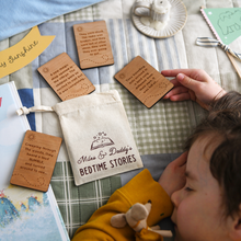 Personalised bedtime story set for dads and children featuring engraved wooden story prompt cards and a drawstring cotton bag reading 'Miles & Daddy’s Bedtime Stories', displayed on a patchwork quilt with a soft toy, picture book, and a young child sleeping beside it.