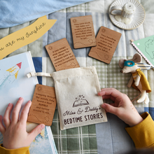 Personalised bedtime story set for dads and children featuring engraved wooden story prompt cards and a drawstring cotton bag reading 'Miles & Daddy’s Bedtime Stories', displayed on a patchwork quilt with a soft toy, picture book, and a young child holding one of the cards.