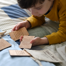 A young child is lying on a bed, covered with a patchwork quilt, reading wooden story prompt cards from a personalised 'Bedtime Stories' bag, designed for storytelling with Daddy.