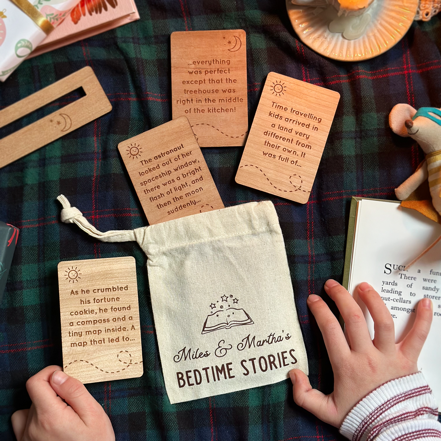 Wooden cards engraved with bedtime story prompts on a plaid fabric, and a personalised 'bedtime stories' cotton bag, with a child's hands interacting with them.