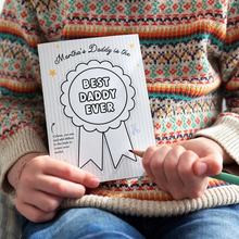 A child wearing a colourful, patterned jumper holds a personalised Father's Day card that reads "Martha's Daddy is the Best Daddy Ever" with a large rosette design ready to be coloured in. The child is holding a blue pencil, preparing to colour the card.