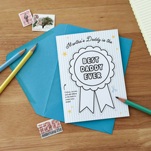 A flat lay of a personalised Father's Day card that reads "Martha's Daddy is the Best Daddy Ever" featuring a large, uncoloured medal design. The card is placed on a wooden table with a bright blue envelope, coloured pencils, and vintage postage stamps nearby.
