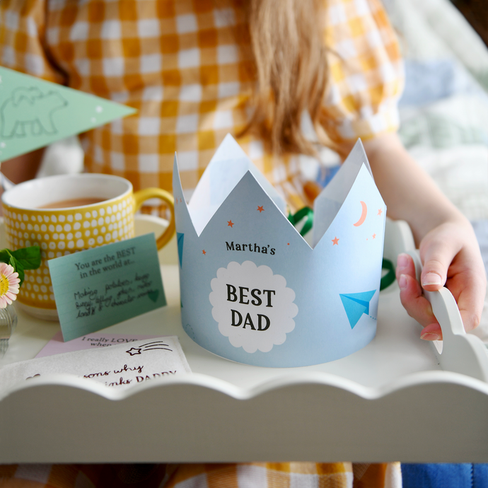 A personalised Best Dad crown with a blue and green paper aeroplane design stands on a Father's Day breakfast tray that is being held by a young child. The wooden tray has a cup of tea, gifts, and keepsakes for Daddy. It's a lovely sentimental scene for Father's Day gifting.