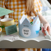 A personalised Best Dad crown with a blue and green paper aeroplane design stands on a Father's Day breakfast tray that is being held by a young child. The wooden tray has a cup of tea, gifts, and keepsakes for Daddy. It's a lovely sentimental scene for Father's Day gifting.