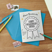 A personalised 'Best Grandad Ever' colour in medal Father's Day card lays on a blue envelope, both on a wooden desk.