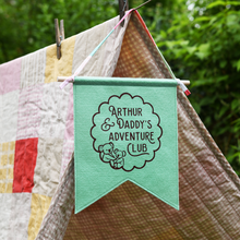A handmade and personalised gentle green felt banner hangs from a wooden dowel and cotton tie at the top of a blanket den in the garden. The flag is personalised with the words Arthur & Daddy's Adventure Club with two little koalas inside a scalloped circle design.