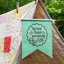 A handmade and personalised gentle green felt banner hangs from a wooden dowel and cotton tie at the top of a blanket den in the garden. The flag is personalised with the words Arthur & Daddy's Adventure Club with two little koalas inside a scalloped circle design.