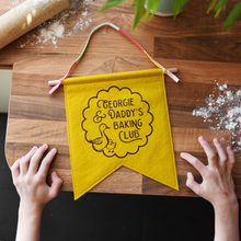 A handmade and personalised felt banner in mustard yellow felt with the words 'Georgie & Daddy's Baking Club' with two little ducks in a scallop design. It has a wooden dowel and cotton tie hanger. The banner lays on a wooden kitchen surface with a young child's hands beside it, and flour sprinkled on the wood.