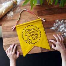 A handmade and personalised felt banner in mustard yellow felt with the words 'Georgie & Daddy's Baking Club' with two little ducks in a scallop design. It has a wooden dowel and cotton tie hanger. The banner lays on a wooden kitchen surface with a young child's hands beside it, and flour sprinkled on the wood.