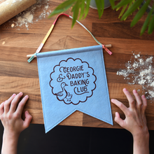 A handmade and personalised felt banner in soft blue felt with the words 'Georgie & Daddy's Baking Club' with two little ducks in a scallop design. It has a wooden dowel and cotton tie hanger. The banner lays on a wooden kitchen surface with a young child's hands beside it, and flour sprinkled on the wood.