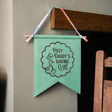 A handmade and personalised felt banner in gentle green felt with the words 'Posy & Daddy's Baking Club' with two little ducks in a scallop design. It has a wooden dowel and cotton tie hanger. The banner hangs from wooden mantle beside a rocking chair.