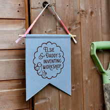 Blue banner with 'Elsie & Daddy's Inventing Workshop' text hanging on a wooden wall.