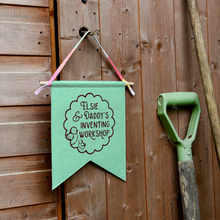 A personalised and handmade felt pennant flag with a wooden hanger and cotton tie, hangs on a shed door. The gentle green flag is personalised with the words Elsie & Daddy's Inventing Workshop, with two birds, inside a scalloped circle design.