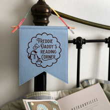 A handmade and personalised felt banner flag in soft blue is hanging from a bedhead. The design on the flag shows the words Freddie & Daddy's Reading Corner with two little penguins inside a scalloped circle pattern.