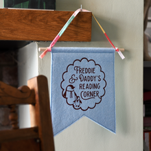A handmade and personalised felt banner flag in soft blue is hanging from a wooden mantle beside a rocking chair. The design on the flag shows the words Freddie & Daddy's Reading Corner with two little penguins inside a scalloped circle pattern.