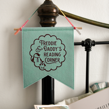 A handmade and personalised felt banner flag in gentle green is hanging from a bedhead. The design on the flag shows the words Freddie & Daddy's Reading Corner with two little penguins inside a scalloped circle pattern.