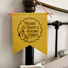 A handmade and personalised felt banner flag in mustard yellow is hanging from a bedhead. The design on the flag shows the words Freddie & Daddy's Reading Corner with two little penguins inside a scalloped circle pattern.