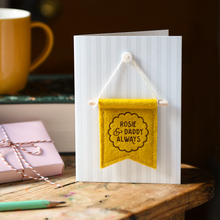 Handmade Father’s Day card with personalised mustard felt banner “Rosie & Daddy Always”, miniature hanging flag keepsake on wooden table. The flag is hung on the card from a tiny mother of pearl button. Beside the card is a book, a mug, a wrapped gift, and a pencil.