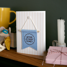 Personalised Father’s Day keepsake card with blue felt banner reading “Arthur & Daddy Always”, handmade mini flag with wooden dowel and button detail. The card stands on a wooden mantle beside a mug, wrapped gift box, candle, and tiny mouse toy.
