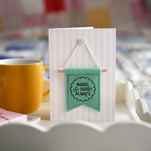 Personalised Daddy and me keepsake card with green felt banner “Mabel & Daddy Always”, handmade Father’s Day gift with stitched flag detail. The banner has a wooden dowel and is hung on the card from a tiny mother of pearl button. The card stands on a wooden breakfast tray beside a mug and wrapped gift.