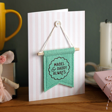 Personalised Daddy and me keepsake card with green felt banner “Mabel & Daddy Always”, handmade Father’s Day gift with stitched flag detail. The banner has a wooden dowel and is hung on the card from a tiny mother of pearl button. The card stands on a wooden mantle beside a mug, wrapped gift box, candle, and tiny mouse toy.