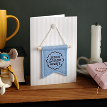 Personalised Father’s Day keepsake card with blue felt banner reading “Arthur & Daddy Always”, handmade mini flag with wooden dowel and button detail. The card stands on a wooden mantle beside a mug, wrapped gift box, candle, and tiny mouse toy.