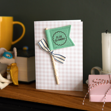 Personalised Father’s Day card with green felt flag reading “Elsie & Daddy”, handmade keepsake card with ribbon and wooden stick detail. The card stands on a wooden mantle beside a book, mug, candle, and wrapped gift.