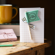 Personalised Father’s Day card with mustard felt flag reading “Henry & Daddy”, handmade keepsake card with ribbon and wooden stick detail. The card stands on a wooden table beside a book, mug, pencil, and wrapped gift.