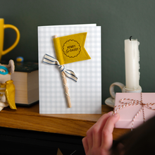 Personalised Father’s Day card with mustard felt flag reading “Henry & Daddy”, handmade keepsake card with ribbon and wooden stick detail. The card stands on a wooden mantle beside a book, mug, candle, and wrapped gift.